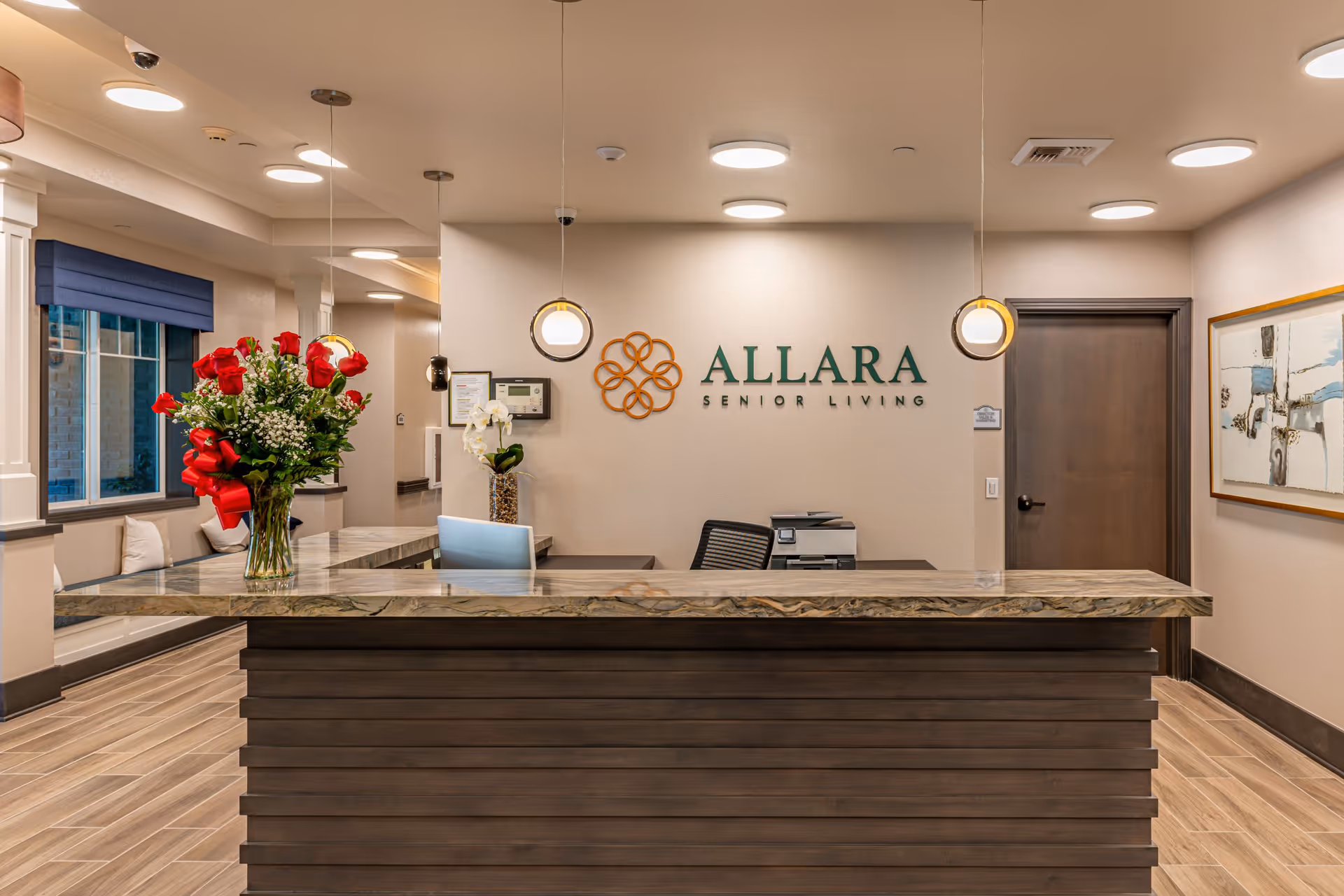 Reception desk area at Allara Senior Living featuring a marble countertop with a vase of red roses, two pendant lights hanging above, a seating area with cushions by a window with a blue blind, and a wall with the Allara Senior Living logo and decorative artwork.