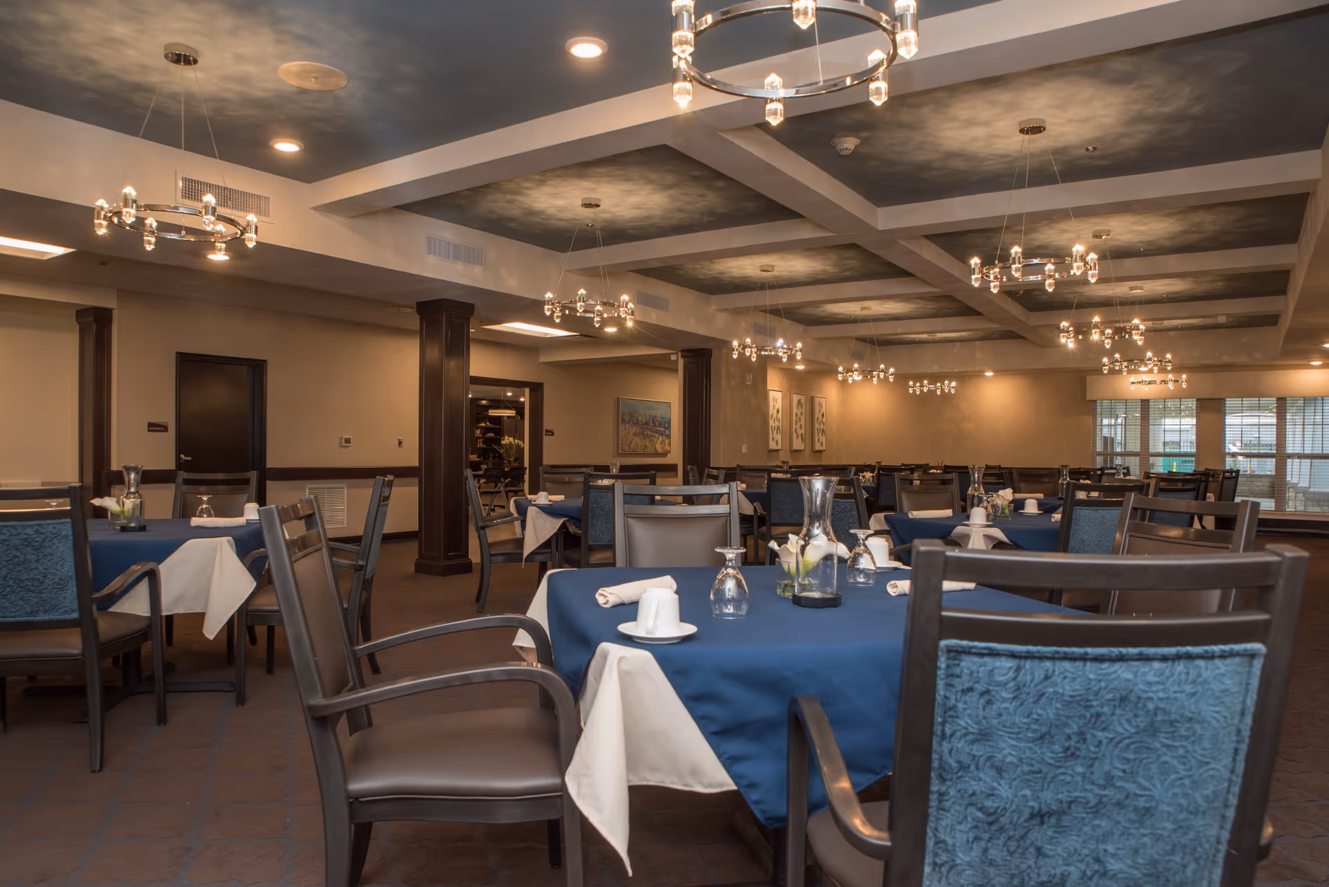 A spacious dining room with multiple tables covered in blue tablecloths and white napkins, set with glassware and small floral centerpieces. The room features elegant chandeliers hanging from a coffered ceiling, dark wood columns, and large windows allowing natural light to enter.