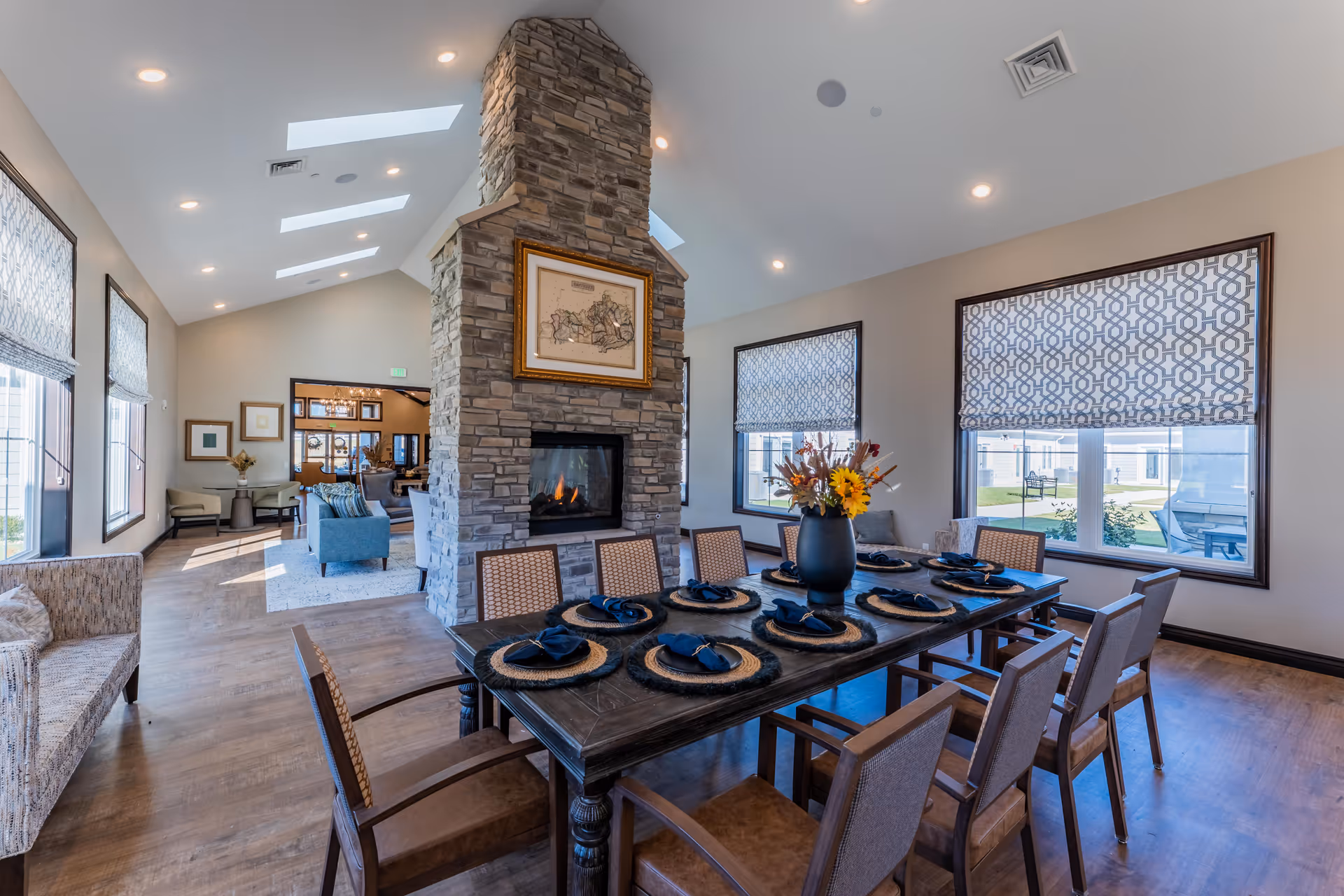 A spacious and well-lit dining area in a senior living facility featuring a long wooden dining table set with eight place settings, each with a woven placemat and a dark napkin. A large stone fireplace with a framed artwork above it divides the dining area from a cozy living room with blue armchairs and a beige sofa. The room has large windows with patterned roller shades, wooden flooring, and skylights in the ceiling allowing natural light to fill the space.