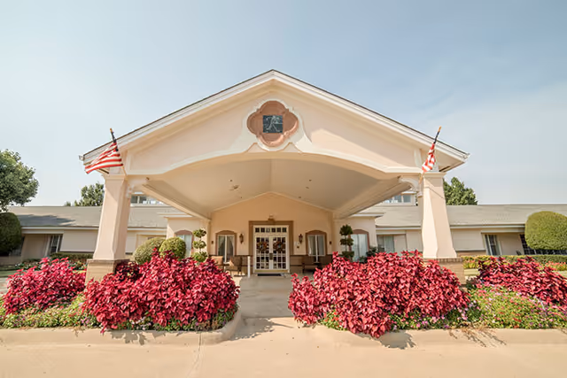 Front exterior view of a single-story building with a covered entrance supported by columns. The entrance is flanked by vibrant red-leafed bushes and two American flags on either side of the roof. The building has a light peach and beige color scheme with a decorative window above the entrance.