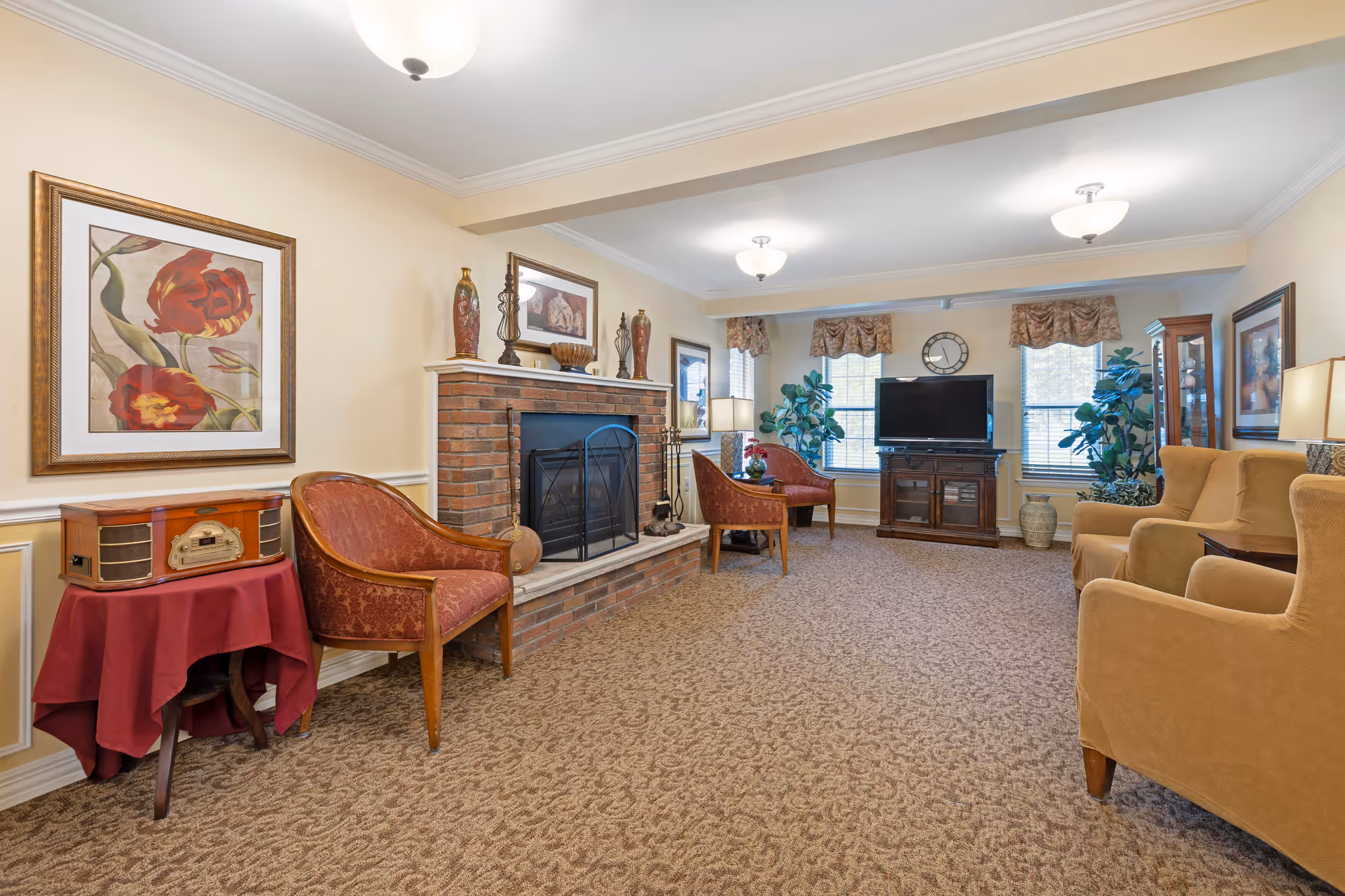 A cozy living room with beige walls and carpet, featuring a brick fireplace with decorative items on the mantel. There are several upholstered chairs and a small table with a vintage-style radio. The room has three windows with valances, a flat-screen TV on a wooden stand, and two large potted plants near the windows.