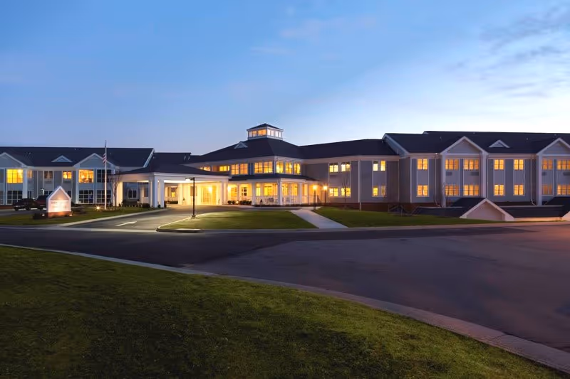 Front exterior of a large illuminated senior living facility at dusk with a circular driveway and lawn.