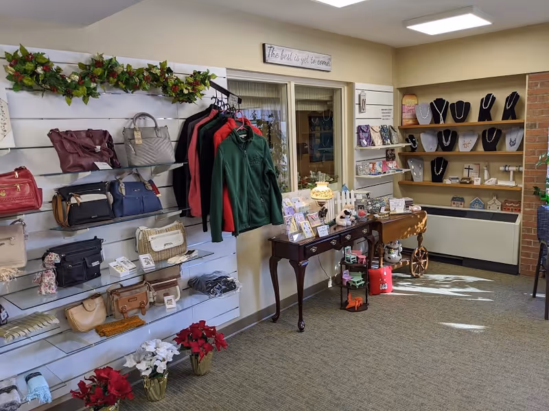 Interior view of a gift shop area in a senior living facility displaying handbags, jackets, jewelry, greeting cards, and decorative items on shelves and tables. There is a sign above the window that reads 'The best is yet to come.'