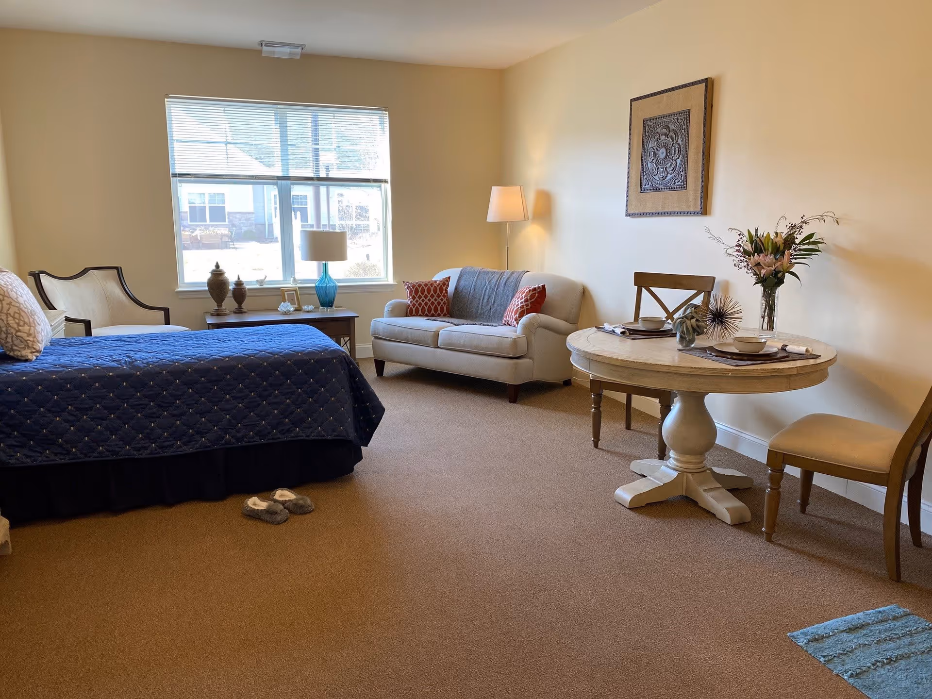 A cozy senior living room with a bed covered in a blue quilt, a beige armchair, a small wooden side table with a blue lamp and decorative items, a beige loveseat with two red patterned pillows, a round wooden dining table set for two with flowers and decorative pieces, and a framed artwork on the wall. A window lets in natural light, and slippers are placed on the carpeted floor.