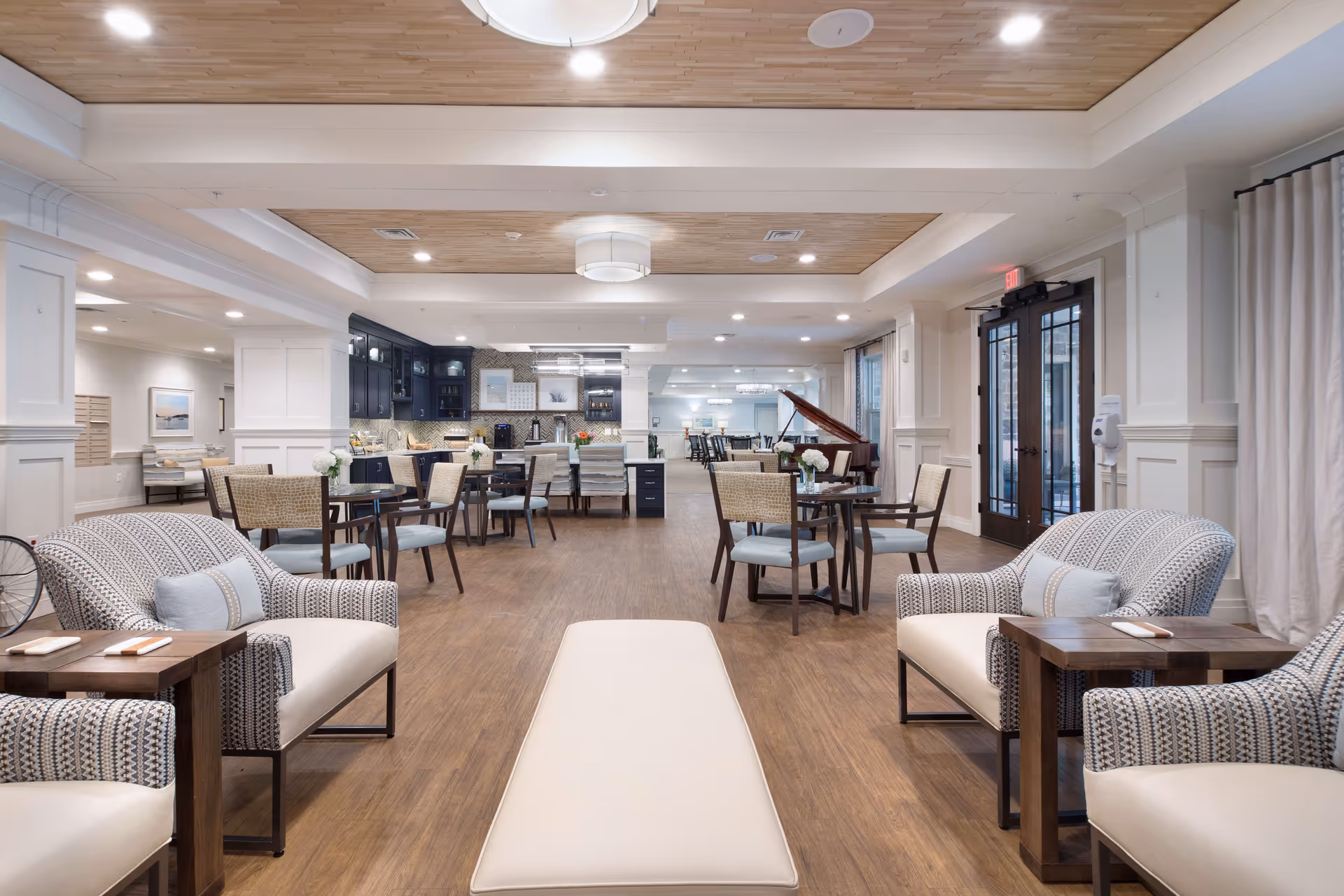 Open, bright communal lounge and dining area with upholstered chairs, tables, and a kitchenette.