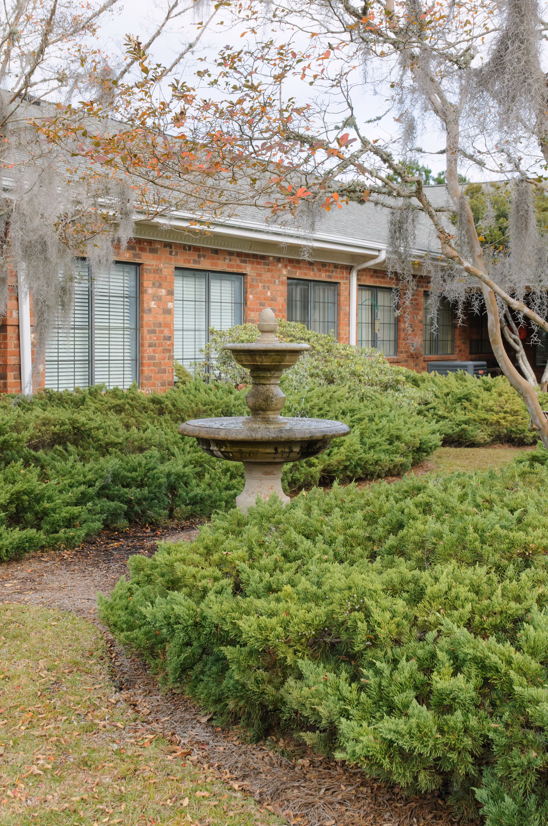 A stone fountain surrounded by green shrubs in front of a brick building with windows.