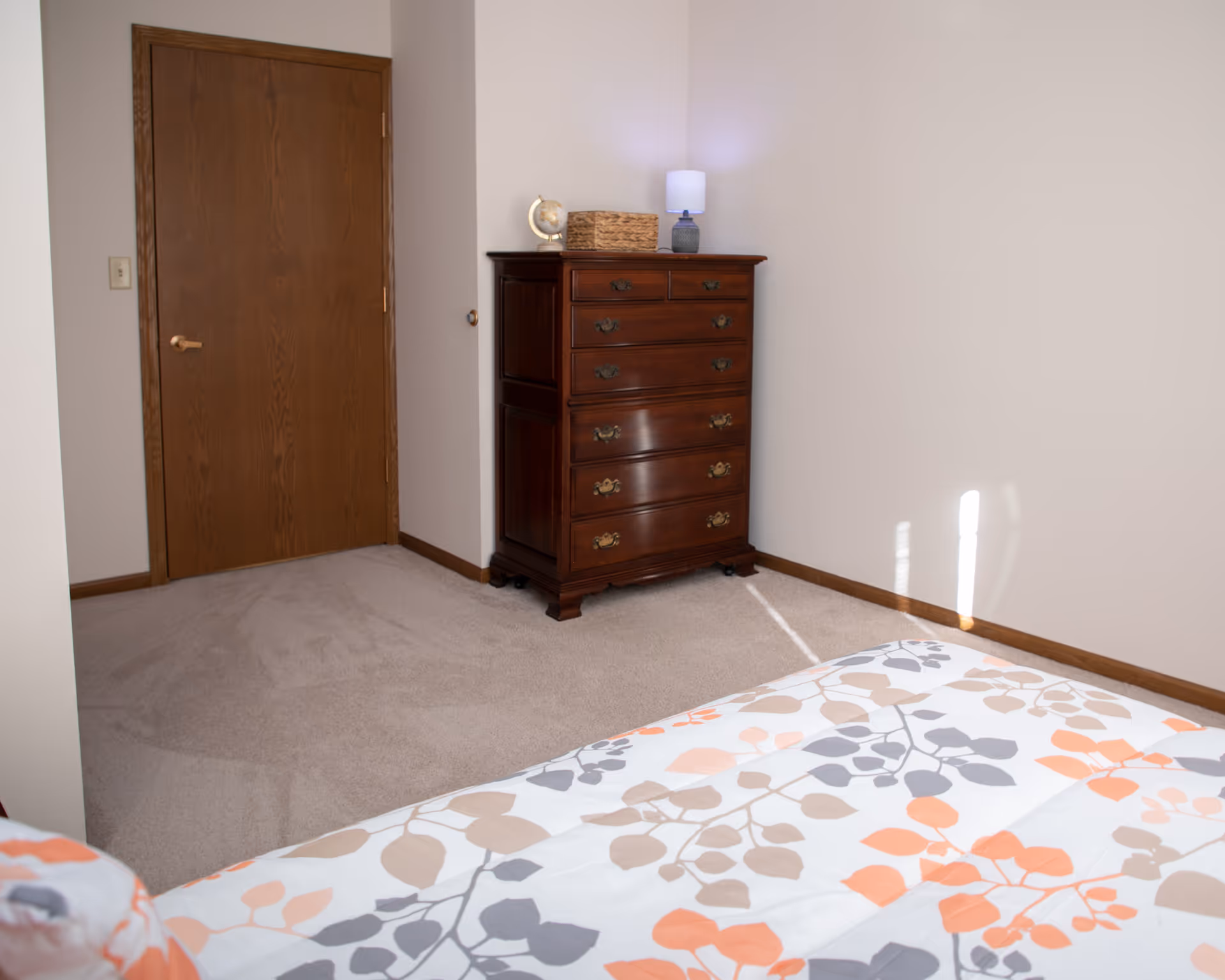 A bedroom corner with a wooden door, a dark wooden chest of drawers topped with a small lamp, a basket, and a decorative globe. Part of a bed with a floral-patterned bedspread in shades of orange, gray, and beige is visible in the foreground.