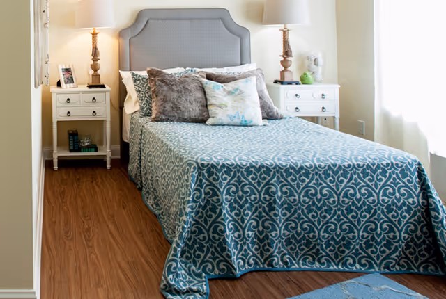 Sunlit bedroom with a patterned blue bedspread, upholstered headboard, decorative pillows, and matching nightstands with lamps.