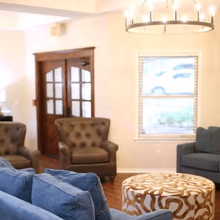 Seating area with blue sofas, tufted gray armchairs, a patterned round ottoman, chandelier, window, and wooden double doors.