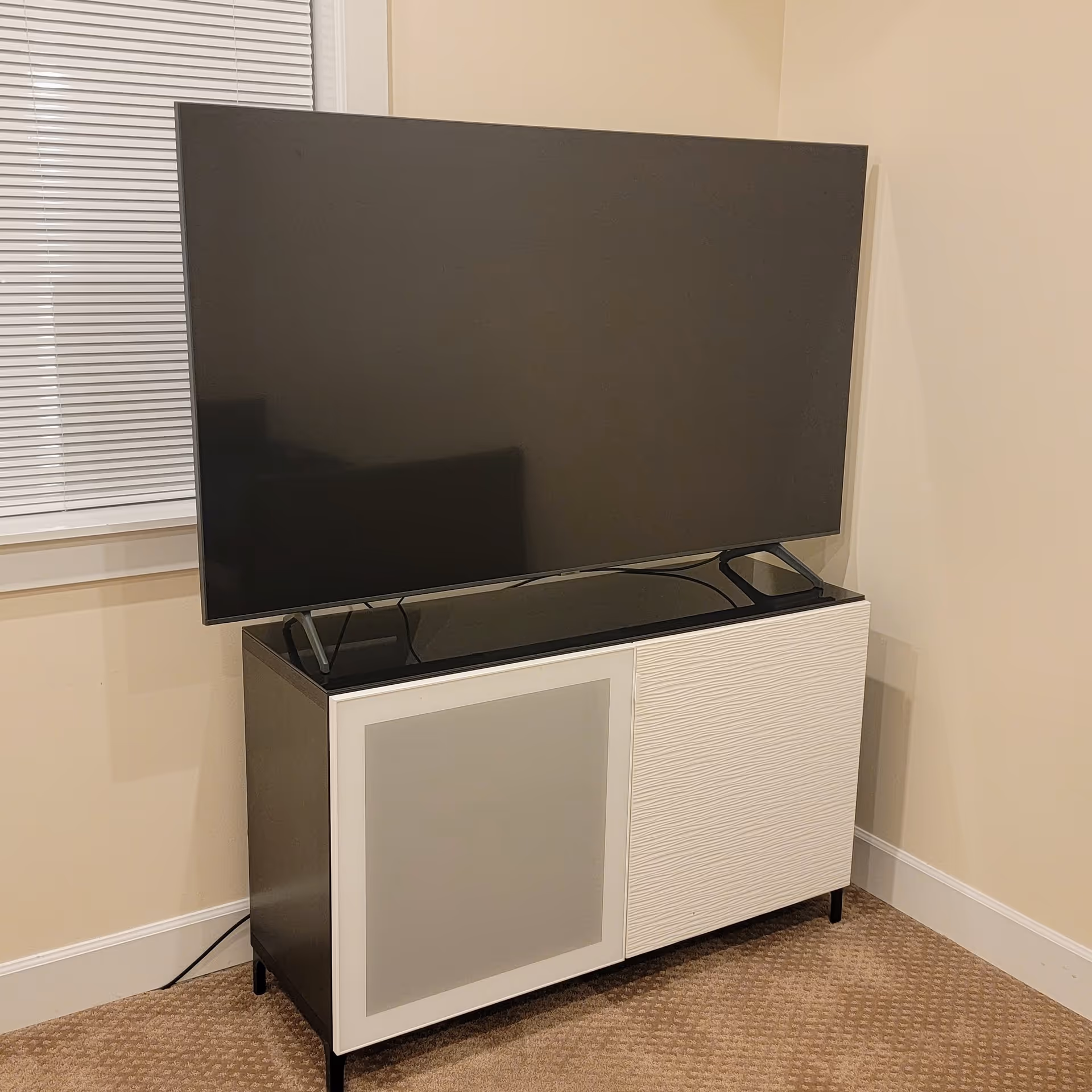 A modern flat-screen television placed on a black and white TV stand with a textured white door and a frosted glass door, positioned against a beige wall next to a window with closed blinds.