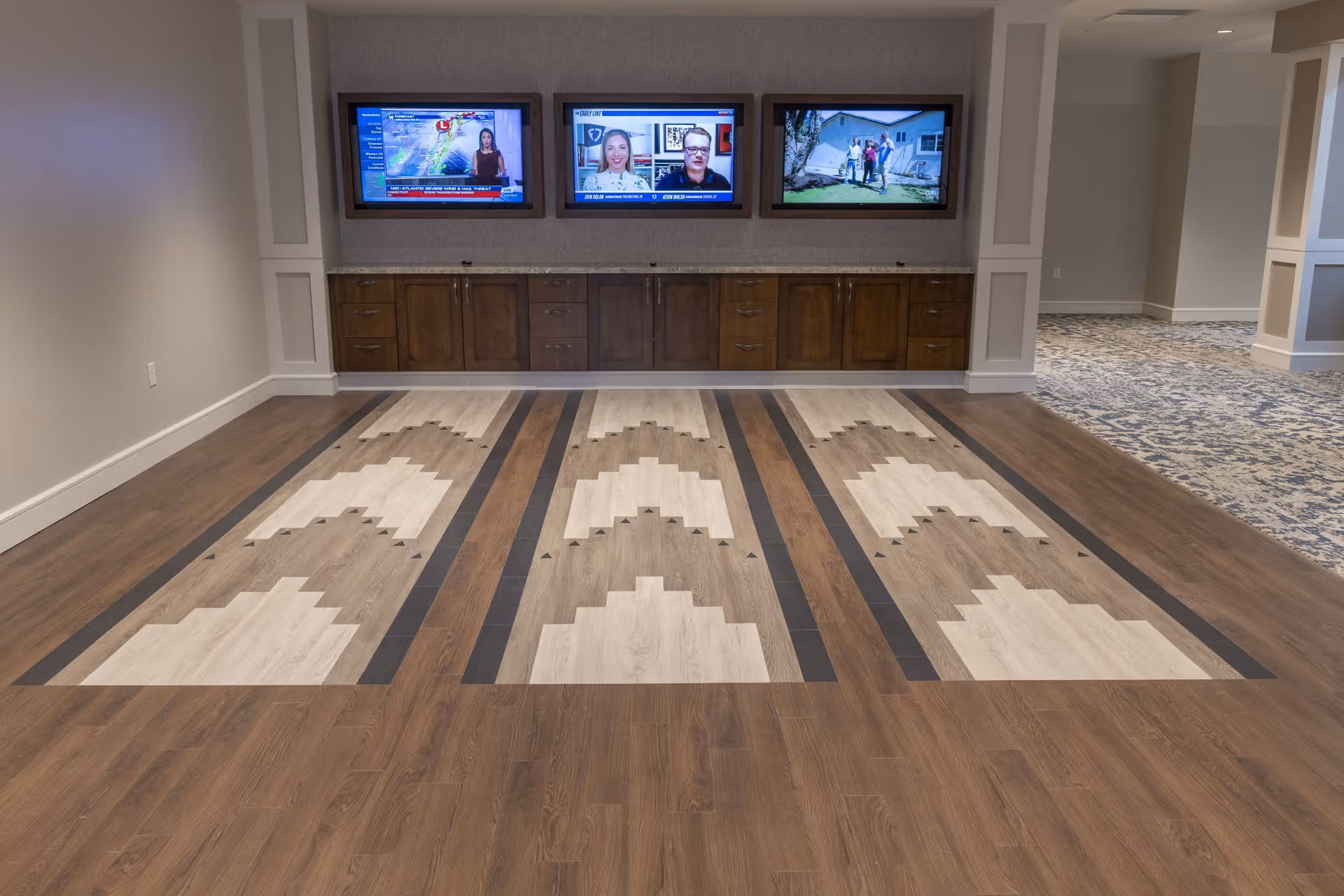 Indoor recreational area with a wooden floor designed for bowling or bocce ball, featuring three lanes with patterned wood inlays. Above the lanes, there are three wall-mounted flat-screen televisions displaying different programs. The area has a cabinet with multiple drawers and doors beneath the TVs, and the surrounding space includes carpeted flooring and neutral-colored walls.