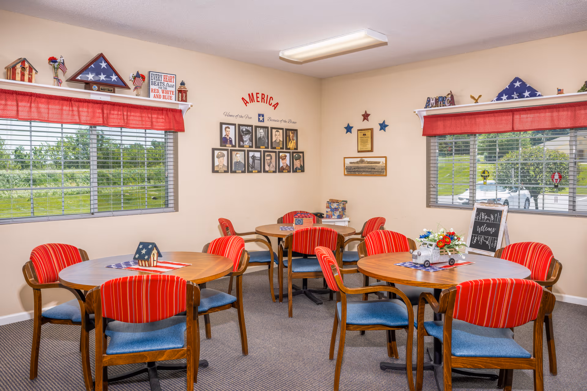 A bright room with three round wooden tables, each surrounded by four chairs with red and blue cushions. The room has two windows with red valances, and patriotic decorations including folded American flags, small flags, and wall art featuring military photos and stars. The walls are beige, and the carpet is gray.