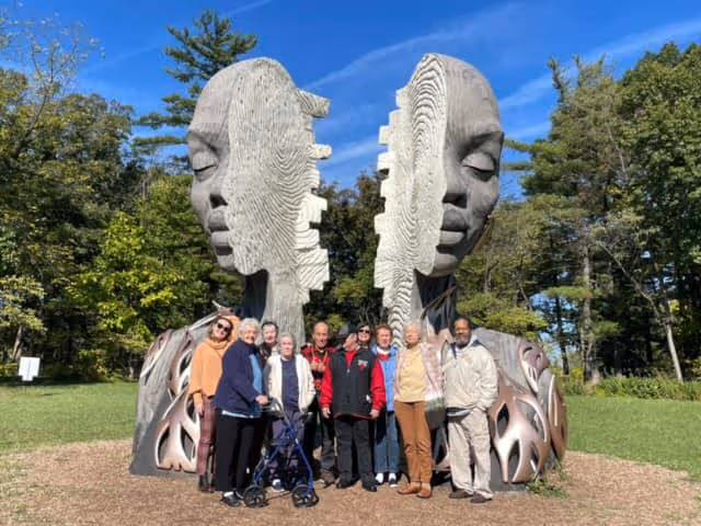 A group of elderly people standing together outdoors in front of a large, artistic sculpture featuring two large, split faces with closed eyes. The setting is a green park area with trees and a clear blue sky.