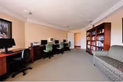 A quiet room with a row of desks each equipped with a computer and office chair along one wall, a large bookshelf filled with books against the opposite wall, and a patterned sofa near the bookshelf. The room has beige walls and carpeted floor with ceiling lights.
