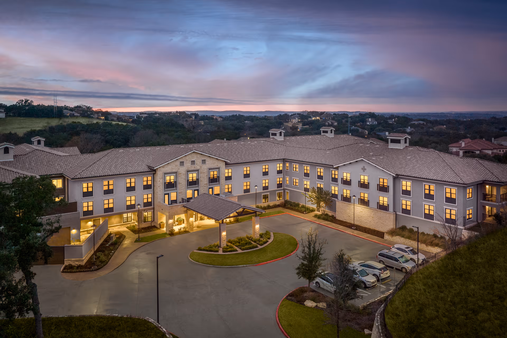Aerial view of Belmont Village Senior Living Lakeway building at dusk with lights glowing from the windows, surrounded by a driveway, parking area with cars, and landscaped greenery under a colorful evening sky.