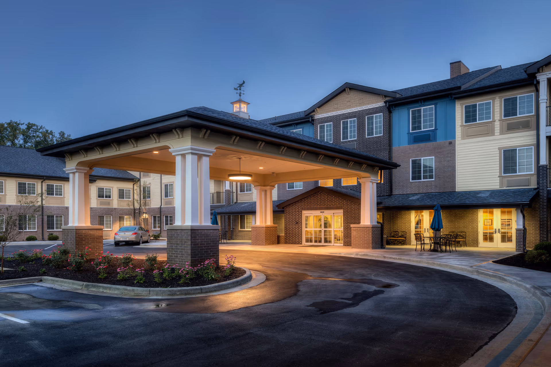 Exterior view of Independence Village of Carmel at dusk, showing a covered entrance with columns, a circular driveway, landscaped flower beds, and a multi-story building with lit windows and outdoor seating area.
