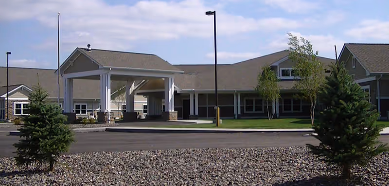 Exterior view of United Pioneer Home building with a covered entrance, surrounded by a paved driveway, small trees, and a landscaped area with rocks under a partly cloudy sky.