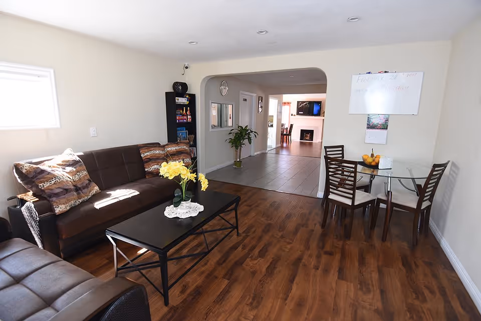 A cozy living room area with brown leather sofas adorned with patterned cushions, a black coffee table with a vase of yellow flowers, and a small glass dining table with four chairs. The room has wooden flooring and light-colored walls. In the background, there is a hallway leading to another room with a fireplace and a TV mounted above it. A whiteboard and a calendar are mounted on the wall near the dining table.