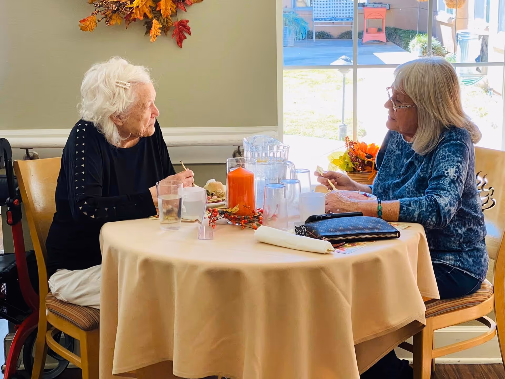 Two elderly women sitting at a round table covered with a beige tablecloth, eating and conversing in a well-lit room with a large window showing an outdoor area. The table has a decorative orange candle centerpiece and several drinking glasses.