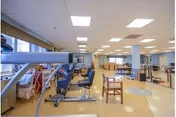 A spacious rehabilitation room with exercise equipment including stationary bikes and parallel bars, chairs, and tables arranged in an open layout with bright overhead lighting and large windows.