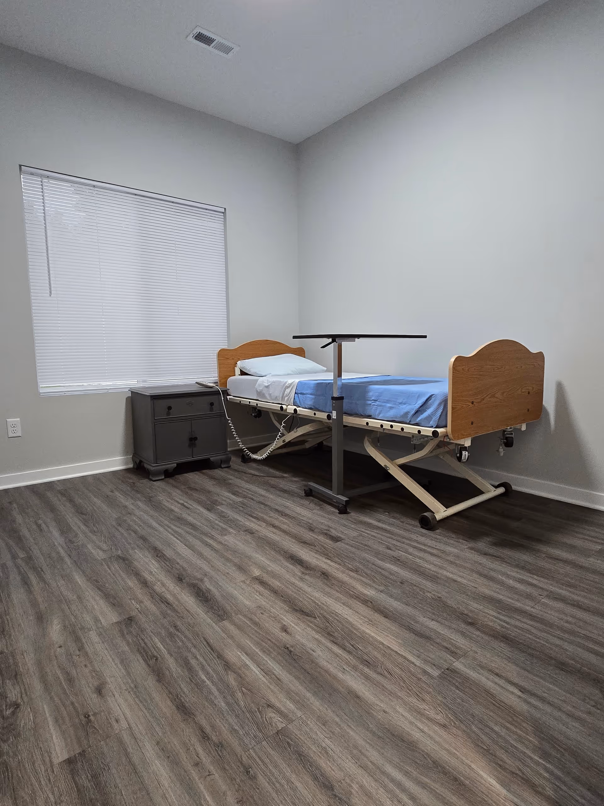 Sparsely furnished bedroom with an adjustable hospital-style bed, overbed table, nightstand, and a window with blinds.