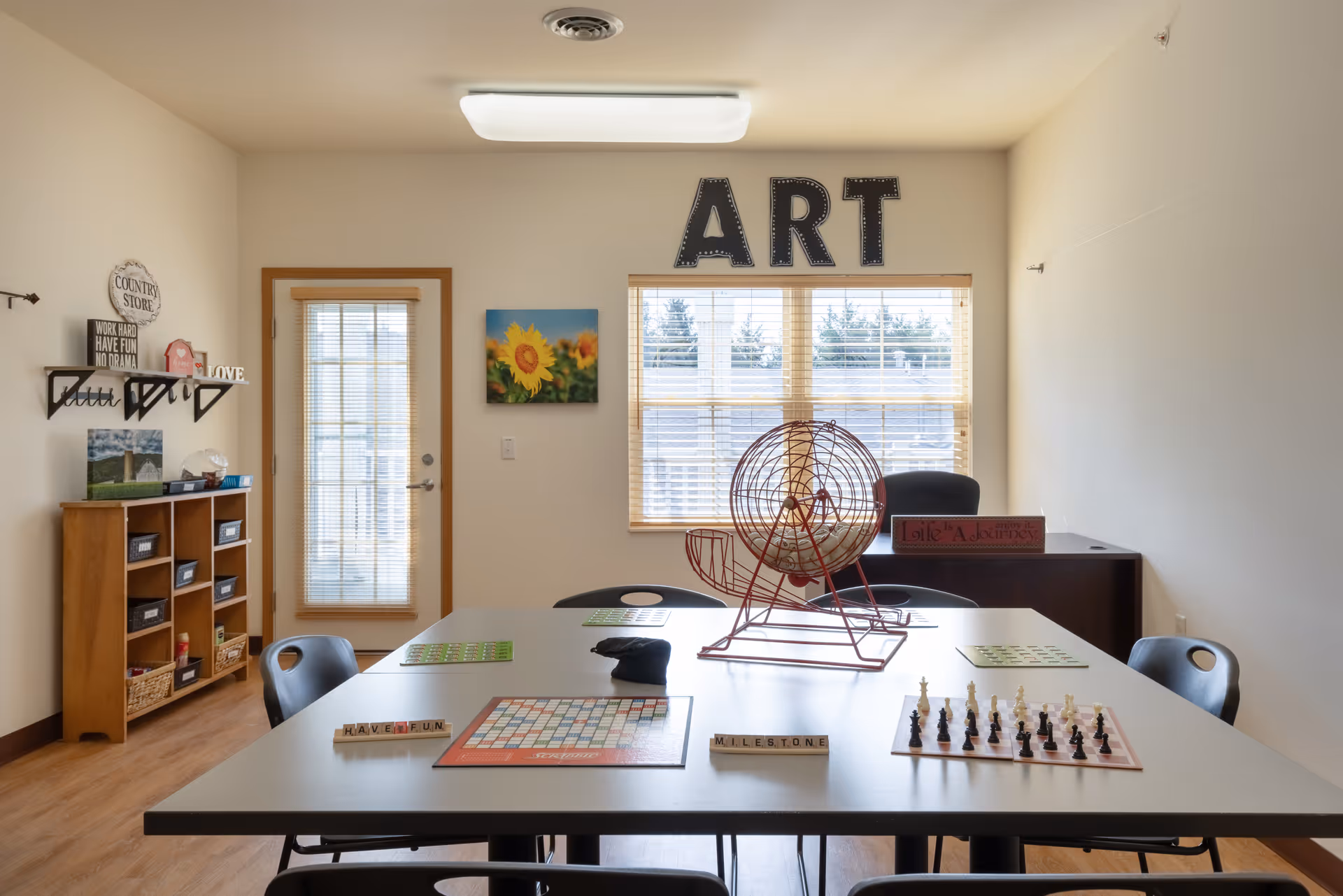 A bright activity room with a large table in the center surrounded by chairs. On the table are a bingo cage, a chessboard, a Scrabble board, and several bingo cards. The wall has a window with blinds and large letters spelling 'ART' above it. There is a door with glass panels and a wooden shelf with baskets and decorative signs on the left side of the room.
