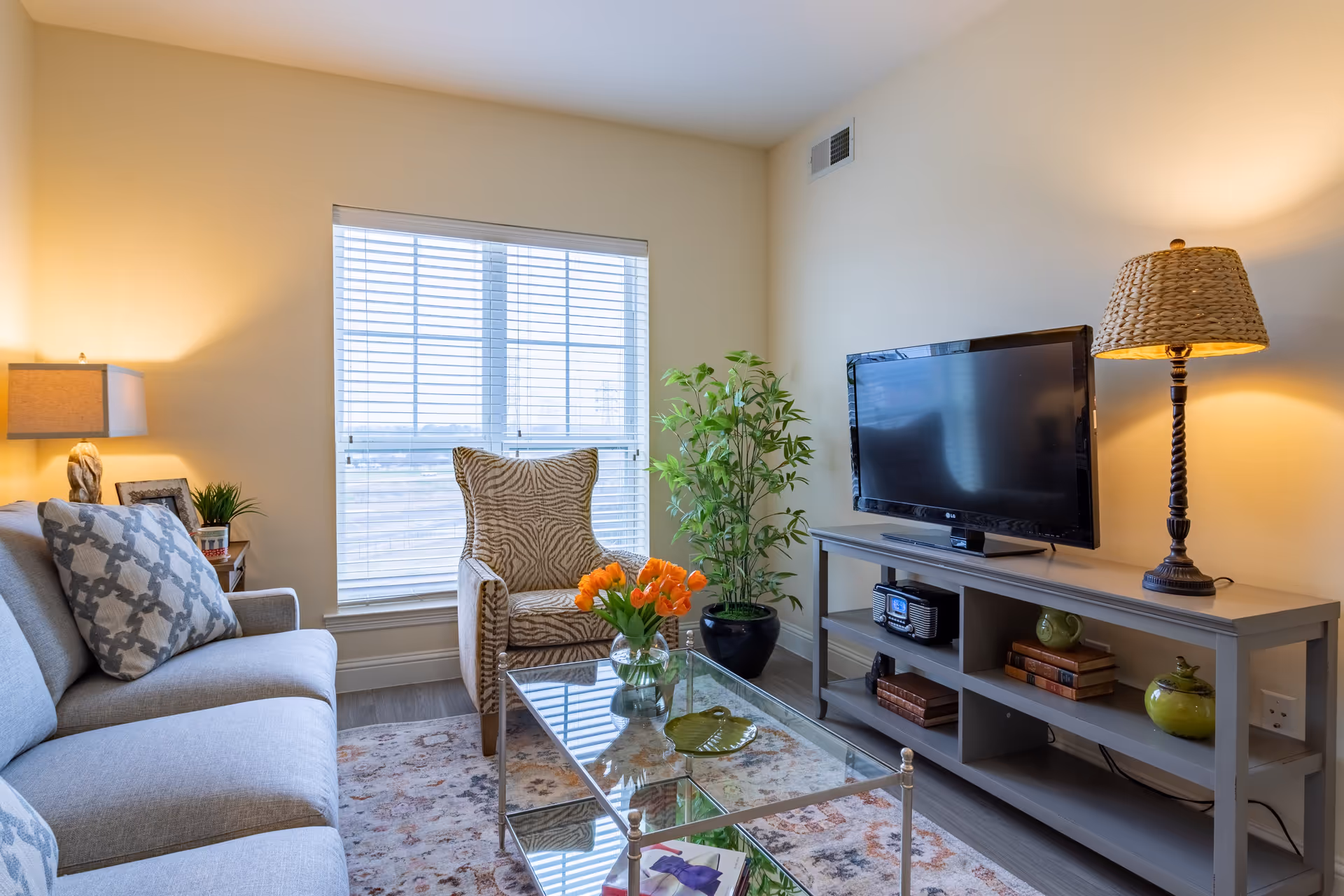 A cozy living room with a gray sofa adorned with patterned pillows, a zebra-print armchair, a glass coffee table with a vase of orange tulips, a TV on a gray console table, a potted plant, and two lamps providing warm lighting.