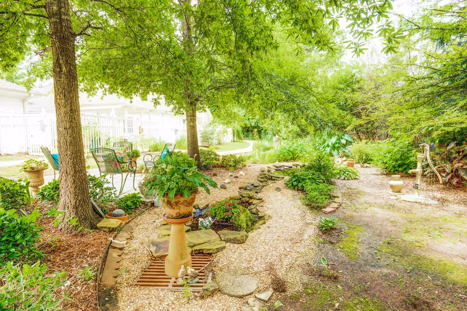 A lush outdoor garden area with green trees providing shade, a gravel pathway bordered by stones, various potted plants, and garden decorations. There are metal chairs and a table with cushions near a white fence in the background.