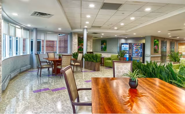 A bright and spacious common area in a senior living facility with multiple wooden tables and cushioned chairs. Large windows on the left side allow natural light to fill the room. There are several green plants placed around the space, and vending machines are visible in the background near a hallway.
