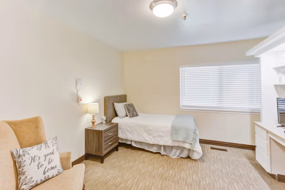 Furnished single bedroom with a bed, nightstand, armchair and window in a senior living unit.