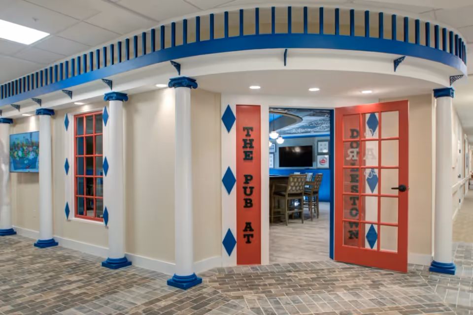 Interior view of an entrance to a pub area named 'The Pub at Doylestown' within a senior living facility. The entrance features white columns with blue bases and capitals, red-framed glass doors with the pub name written vertically, and a tiled floor. Inside, there are high chairs around a table and a TV mounted on the wall.