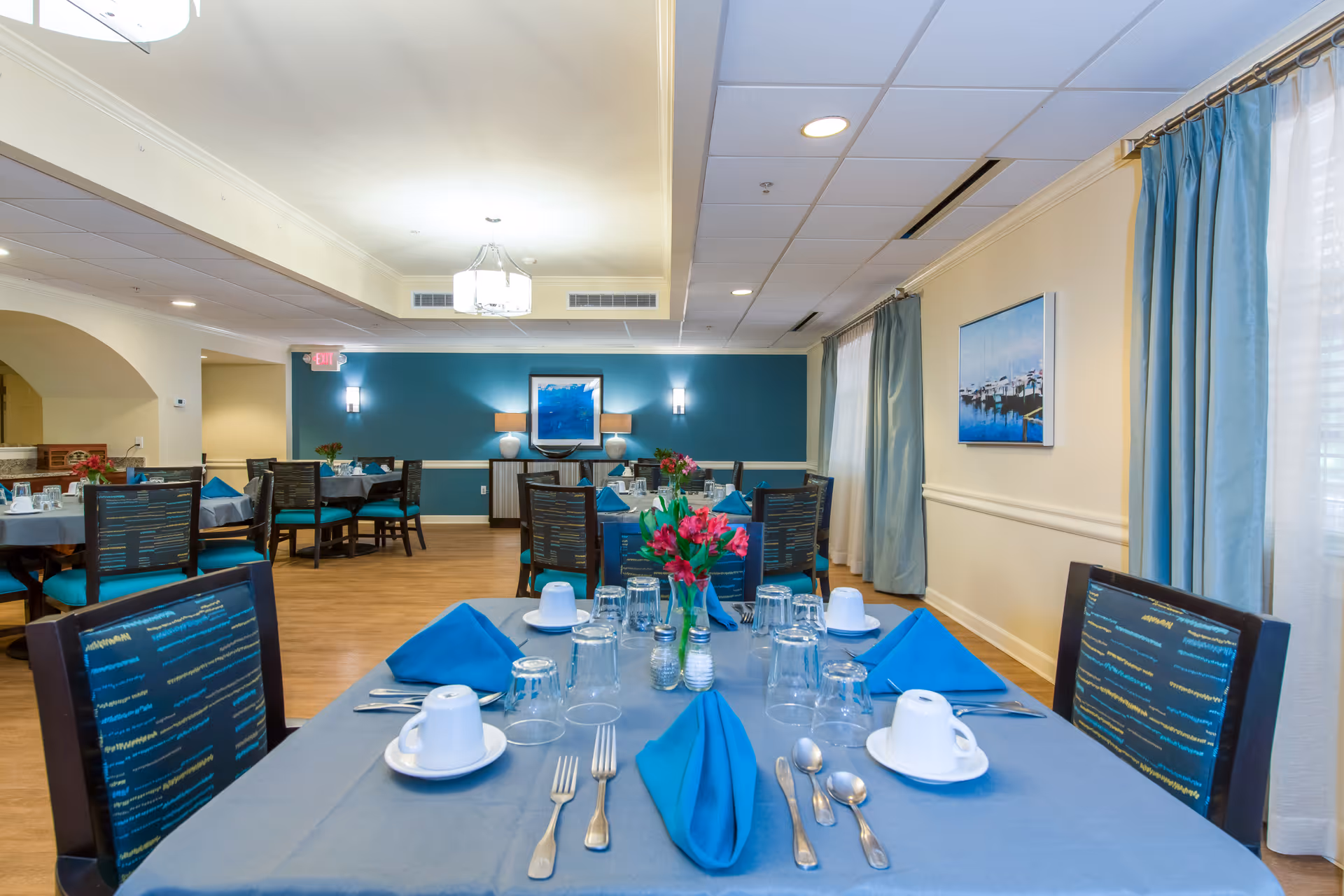 A dining room set up with tables covered in blue tablecloths and matching blue napkins. Each table is set with cups, glasses, silverware, and small flower arrangements. The room has wooden floors, teal accent walls, and large windows with light blue curtains.