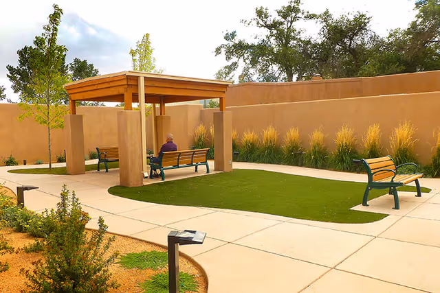 Outdoor courtyard area with a covered wooden pergola, benches, green artificial turf, and surrounding plants and trees. A person is sitting on one of the benches under the pergola.