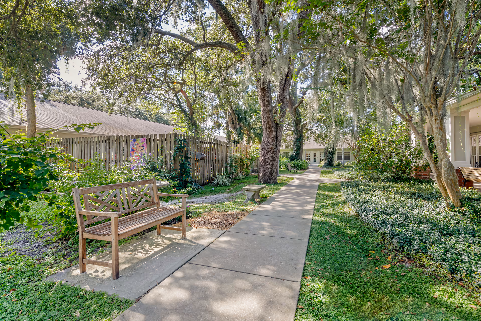 Shaded courtyard walkway with a wooden bench, trees draped in Spanish moss, and building entrances in the background.