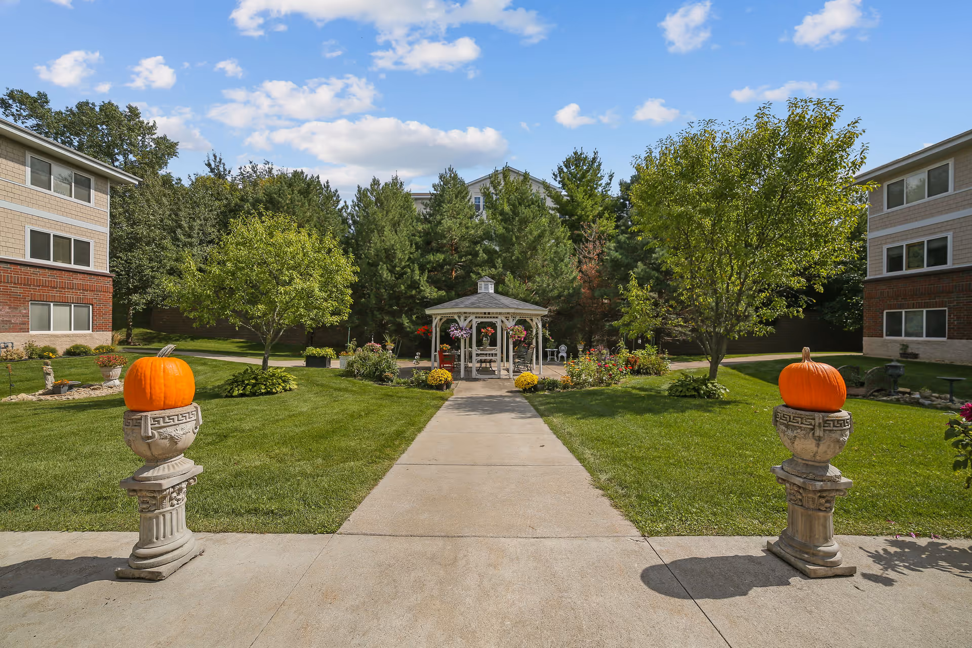 A paved walkway leads to a white gazebo surrounded by green grass, trees, and flower beds. Two large orange pumpkins are placed on decorative stone pedestals on either side of the walkway. Residential buildings with brick and siding are visible on both sides under a partly cloudy blue sky.