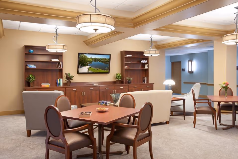 A cozy common area in a senior living facility featuring beige walls and carpet, wooden tables with chairs, two beige armchairs, a beige sofa, wooden bookshelves with plants and books, a flat-screen TV mounted on the wall displaying a scenic lake view, and ceiling lights with fabric shades.
