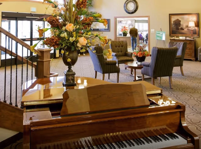 Interior view of a senior living facility lounge area featuring a polished wooden Yamaha piano in the foreground with a large floral arrangement on top. In the background, there are several upholstered chairs arranged around small tables, a clock on the wall, a framed painting, and a person walking away near a doorway.