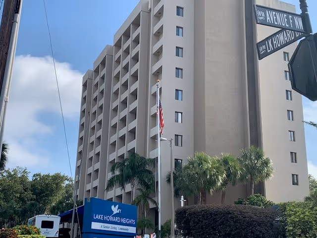 Exterior view of a tall beige senior living facility building with multiple balconies. In front of the building is a blue sign that reads 'Lake Howard Heights, A Senior Living Community' with a white dove logo. There are palm trees and an American flag on a flagpole near the sign. Street signs for '9th Avenue F NW' and 'Lk Howard Dr NW' are visible on the right side of the image under a partly cloudy sky.