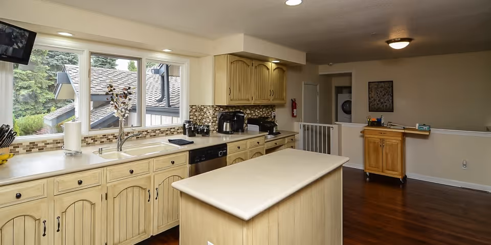 A bright kitchen with wooden cabinets and a central island. The kitchen features a large window overlooking greenery outside, a sink, a dishwasher, and various kitchen appliances on the countertop. The floor is wooden, and there is a small wooden cabinet on wheels in the adjacent room.