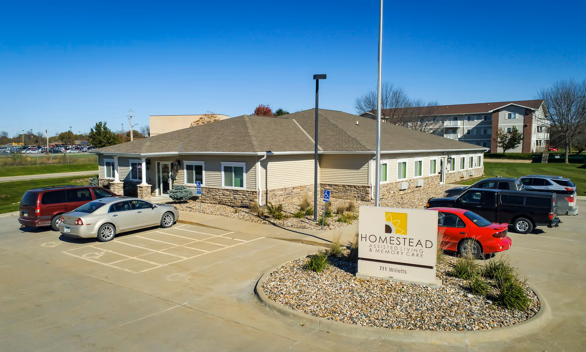Exterior view of Homestead Assisted Living & Memory Care building with a parking lot in front, several parked cars, and a sign displaying the facility name and address 711 Willetts.