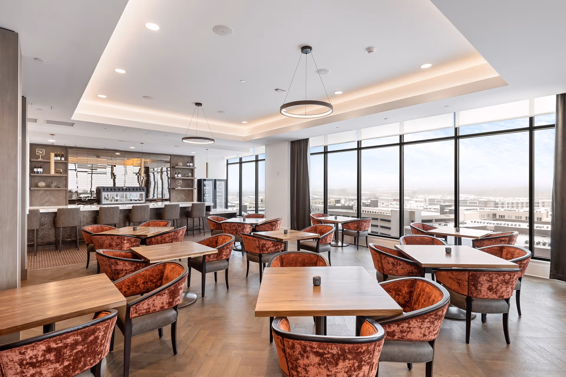 A modern dining room with multiple wooden tables and red velvet upholstered chairs. Large floor-to-ceiling windows provide a panoramic view of the city outside. The room features a bar area with high stools and shelves with bottles and decor. The ceiling has recessed lighting and two circular pendant lights.