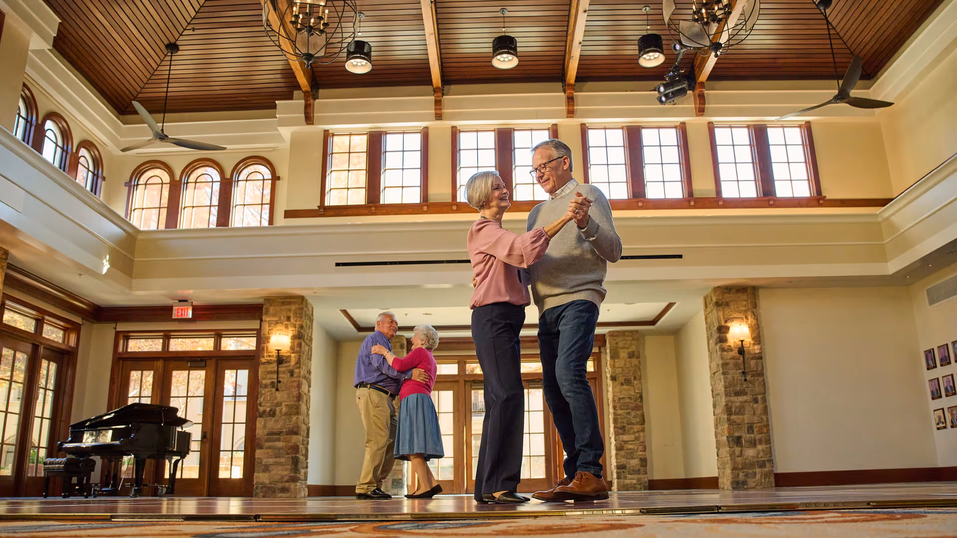 Two elderly couples dancing in a spacious, well-lit room with large windows, wooden ceiling beams, and stone pillars. A grand piano is visible in the background near glass doors.