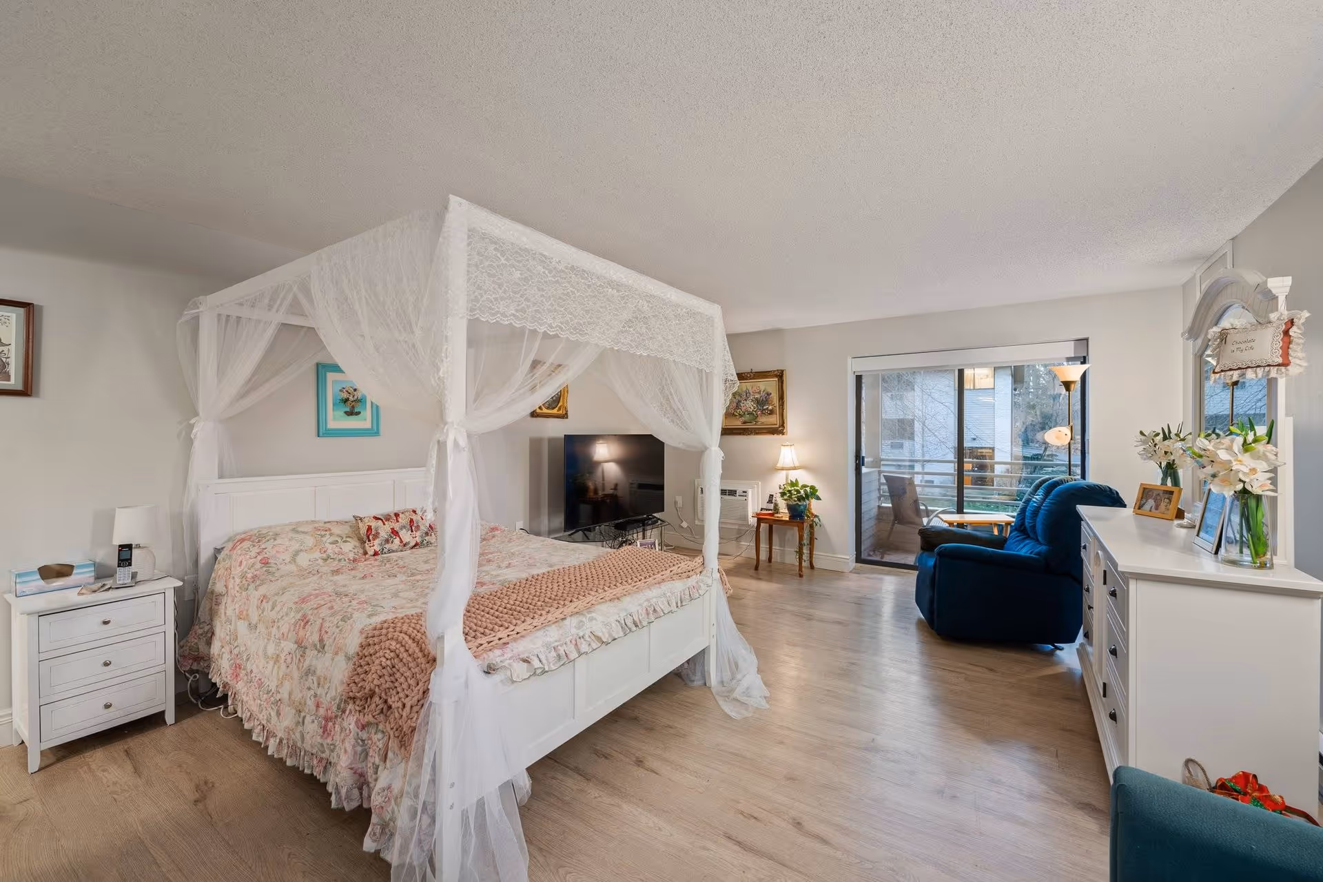 A bright and spacious bedroom featuring a white four-poster bed with sheer lace curtains and floral bedding. The room includes a white nightstand with a lamp and phone, a flat-screen TV on a stand, a small wooden side table with a lamp and plant, a blue recliner chair, and a white dresser with a mirror and decorative items. Sliding glass doors lead to a balcony with outdoor seating, and the room has light wood flooring and neutral-colored walls.