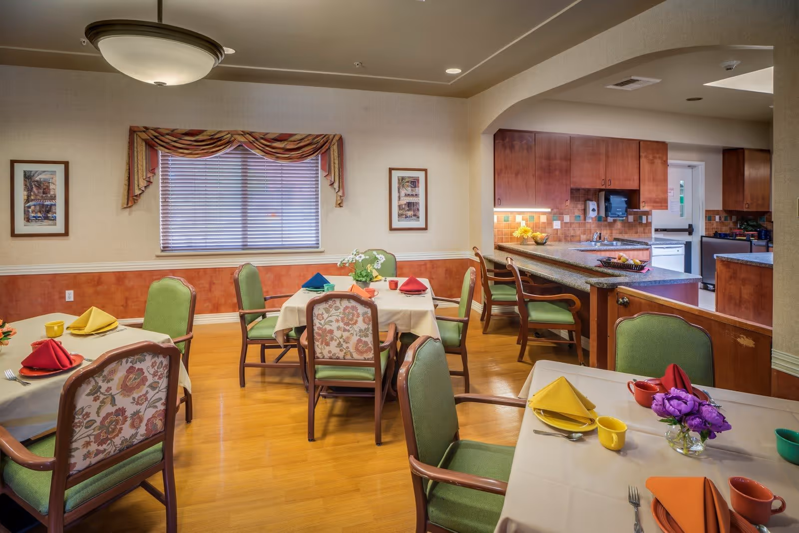 Bright communal dining room with set tables, floral-backed and green chairs, and an open kitchen area.