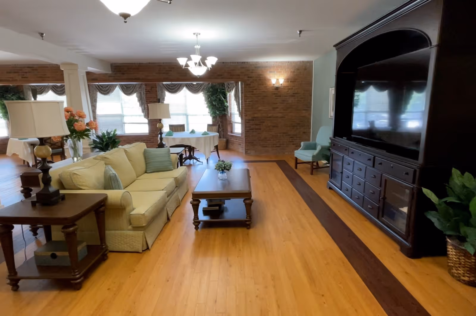 A spacious living room with a light yellow sofa, wooden coffee table with a small flower vase, side tables with lamps, a large dark wood entertainment center with a TV, and a light blue armchair. The room has wooden flooring, brick accent walls, large windows with curtains, and several plants.