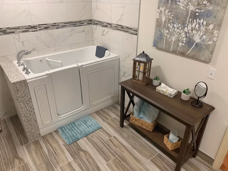 A walk-in bathtub with a door for easy access, surrounded by marble-patterned tiles and a small blue bath mat in front. Next to the tub is a wooden console table holding a decorative lantern, two small potted plants, a box of tissues, a small round mirror, and two wicker baskets with towels. A floral artwork hangs on the wall above the table.