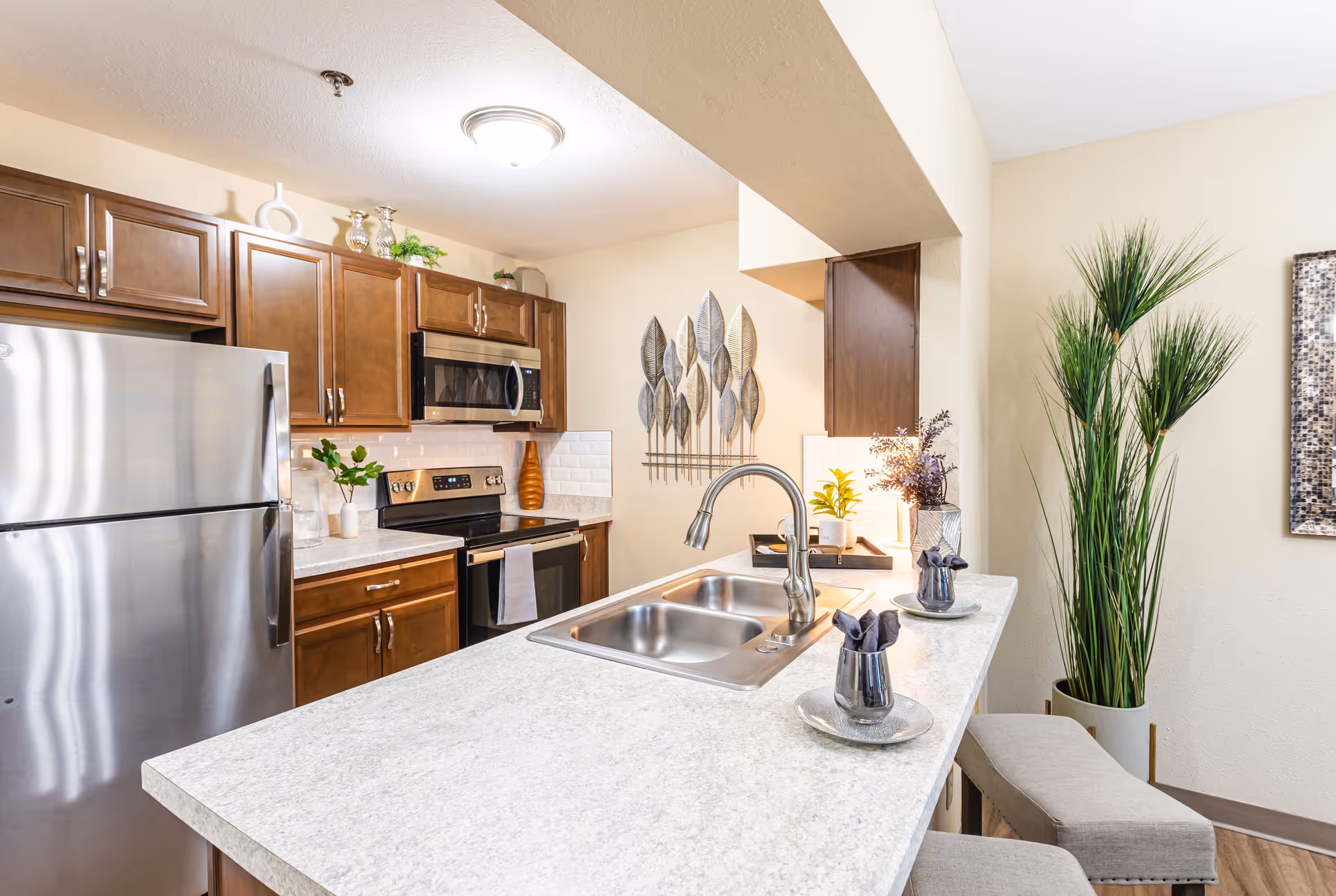 Modern kitchen with stainless steel refrigerator, stove, and microwave. The kitchen features wooden cabinets, a white countertop with a double sink, and decorative plants and items on the counter and walls. There are two cushioned stools at the counter and a tall green plant in the corner.
