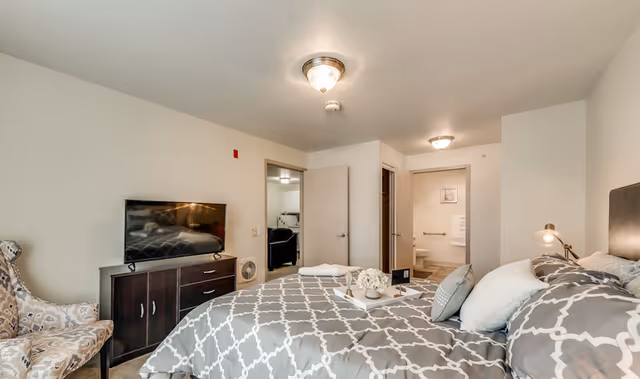 Cozy bedroom with a patterned bedspread, bedside lamp, armchair, television on a dresser, and an open doorway leading to a bathroom.