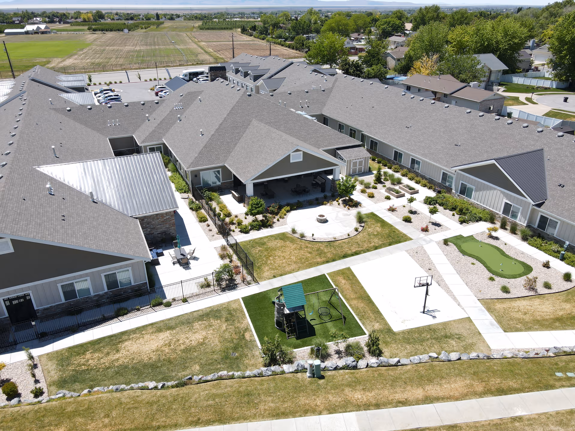 Aerial view of Whisper Cove Assisted Living and Memory Care facility showing a large building with multiple wings surrounding a landscaped courtyard. The courtyard features a covered patio area with seating, a fire pit, a small playground, a basketball hoop, and a putting green. The surrounding area includes parking spaces, residential homes, and open fields in the distance.