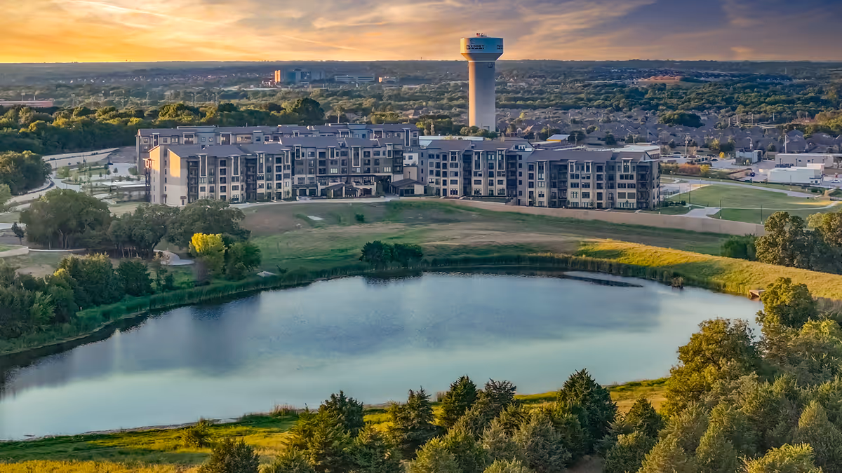 Aerial view of a senior living facility named Touchmark at Emerald Lake, showing a large multi-story building beside a pond with surrounding greenery and trees under a sunset sky.